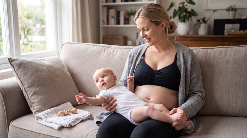 A calm young mother sits on a sofa by a bright window in a lived-in living room, holding her wide-awake four-month-old baby who has unlatched and looks curiously around the room; she wears a dry black wireless nursing bra under an open cardigan